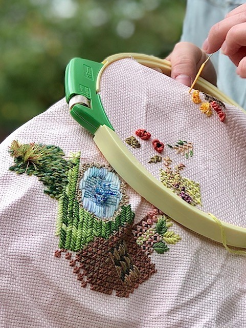 A creamy colored fabric already partially embroidered in a number of colors and stitches, and an embroidery hoop in place as a hand adds more stitches.