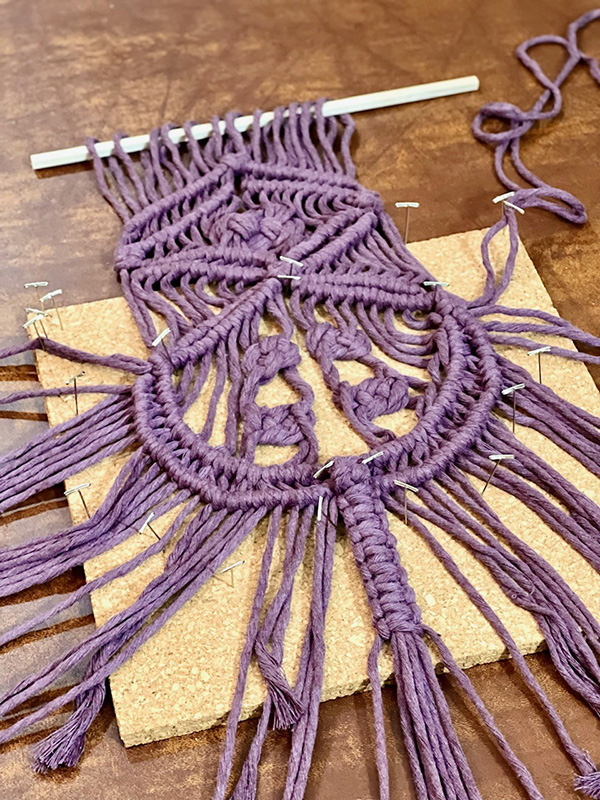A complex macrame hanging done in soft purple yarn pinned flat on a cork square to display the work in progress.