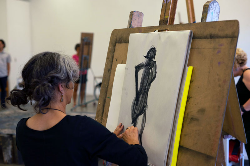 A woman sketching with charcoal on a large piece of paper clipped to an easel.