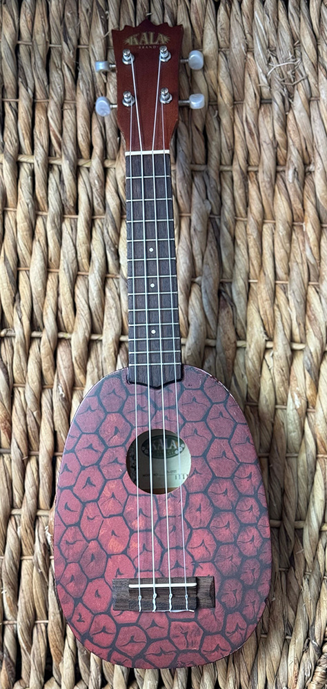 A ukelele with a pineapple pattern painted on the body laying on a rush mat.