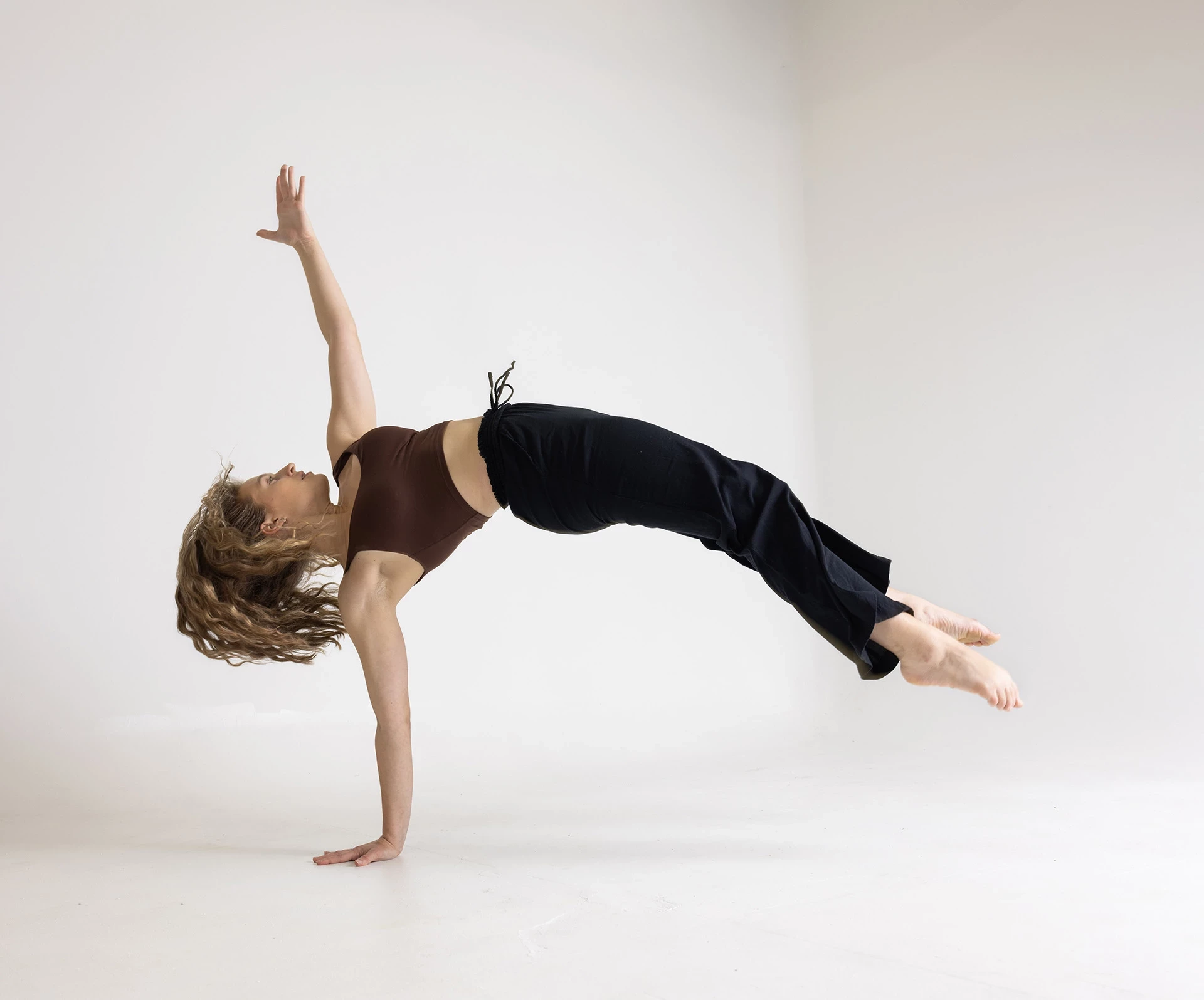 Dancer in a white studio lifting herself up on one hand, body parallel with the floor, other hand reaching to the sky.
