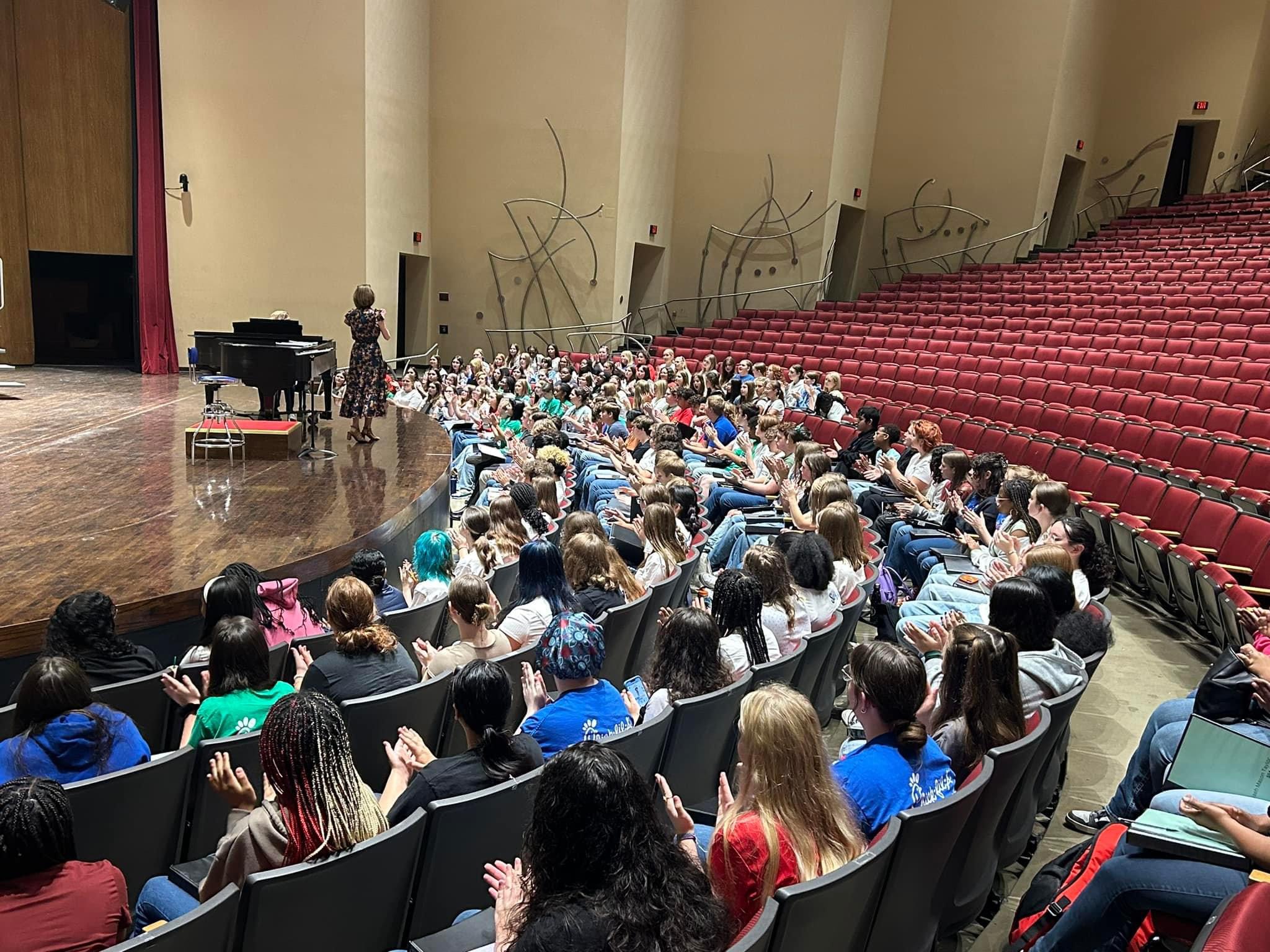 Photo taken from the recital hall seats facing the stage. A diverse and colorfully dressed body of student singers fill the first 4 rows of seats while director Beth Wilson speaks next to a concert piano on stage.