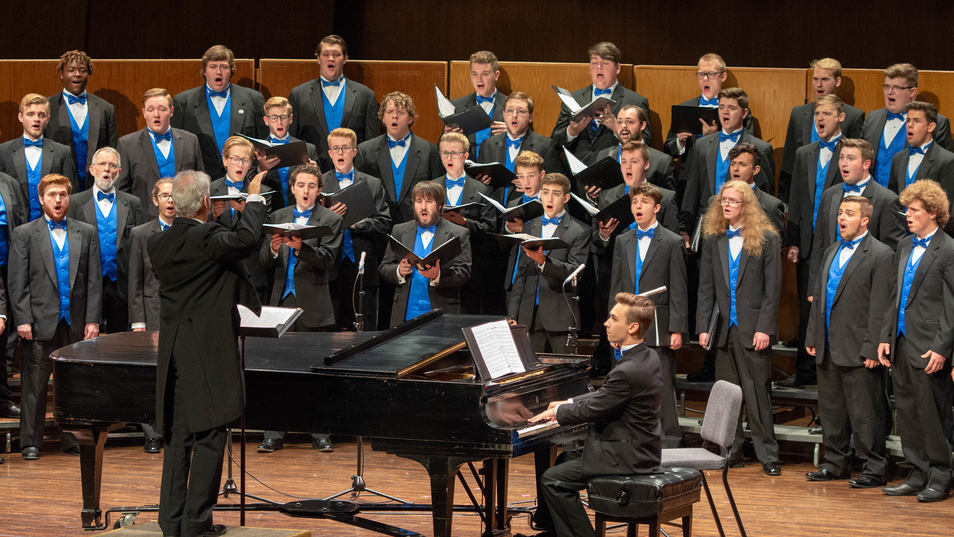 University of Kentucky Men's Choir, Dr. Jefferson Johnson, Director​​, the choir stands facing the camera onstage at the Singletary Center wearing performance black tux, with Dr. Johnson conducting in the foreground next to an accompanist playing a grand piano