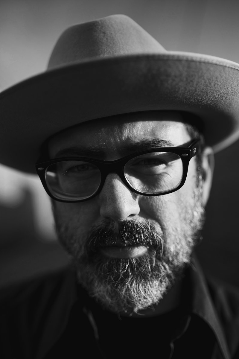 Black and white close up portrait of Brett Ratliff, bearded, with dark eye glasses and light fedora hat