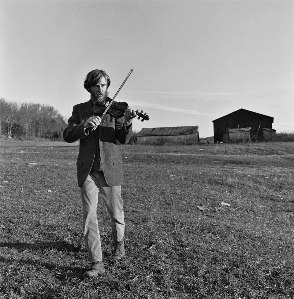 Black and white photograph of John Harrod playing violin wearing a dark blazer and light pants, in a field with a few barns in the background