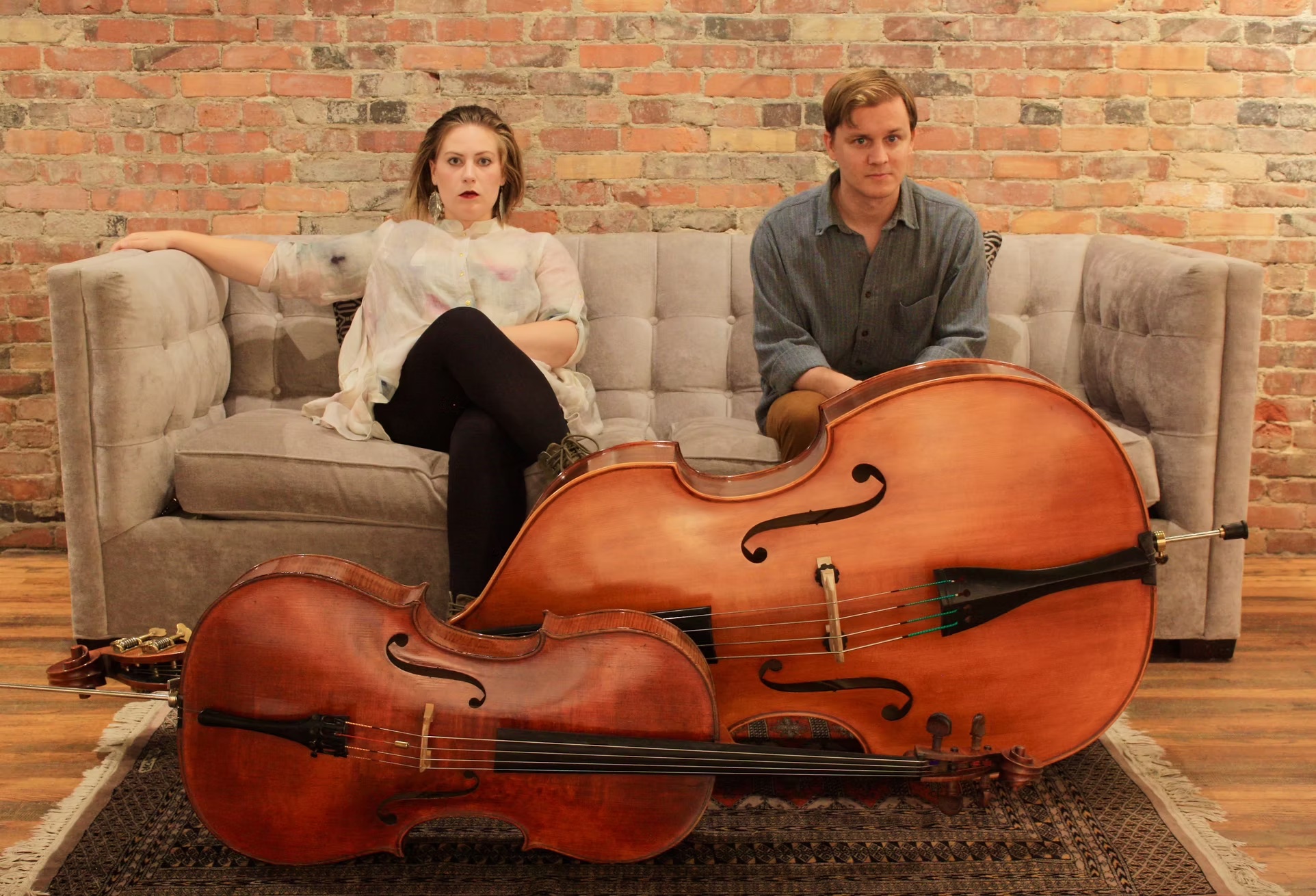 Image of Lindsey Paige Miller and Scott Mitchell Gorski sitting on large tan sofa, against a brick wall background, with a cello and upright bass in front of them.