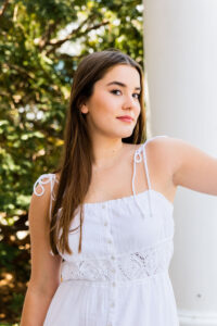 Image of Ella Webster in white dress against a large white column and green leaves in the background