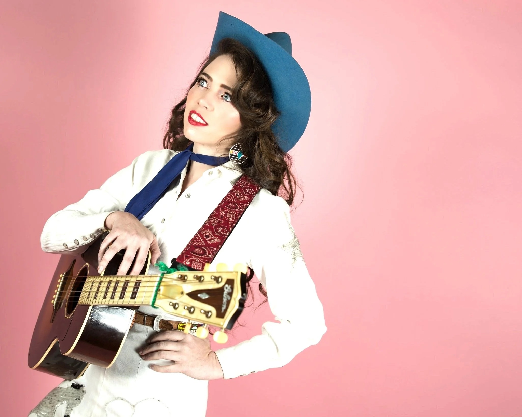 Image of Martha Spencer in white cowgirl outfit, blue cowboy had, holding a guitar, against a pink background