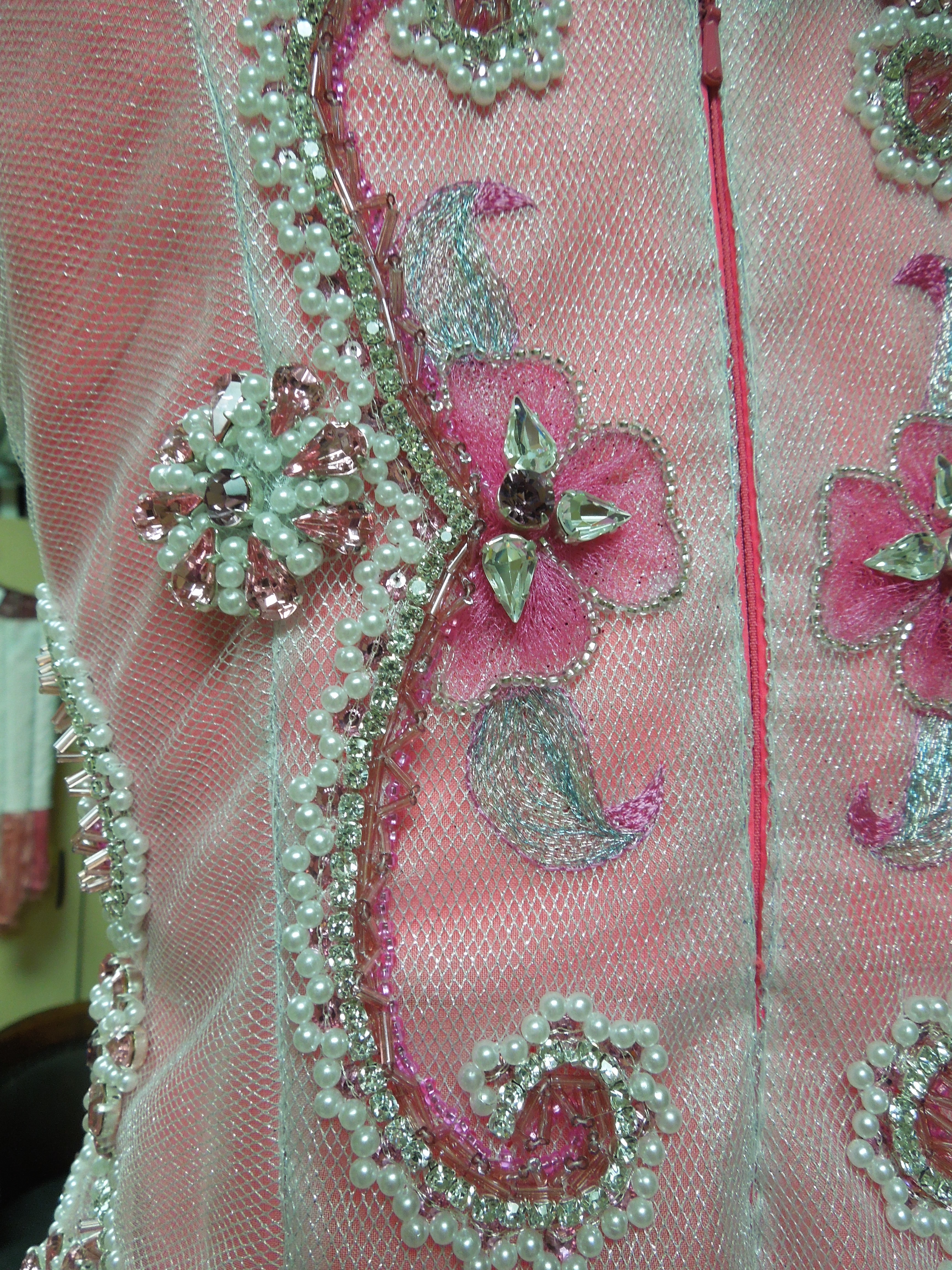 Pearls, seafom beads, and faux gems adorn a sheer metallic fabric layered over the vibrant pink bodice of a dress on a manequin.
