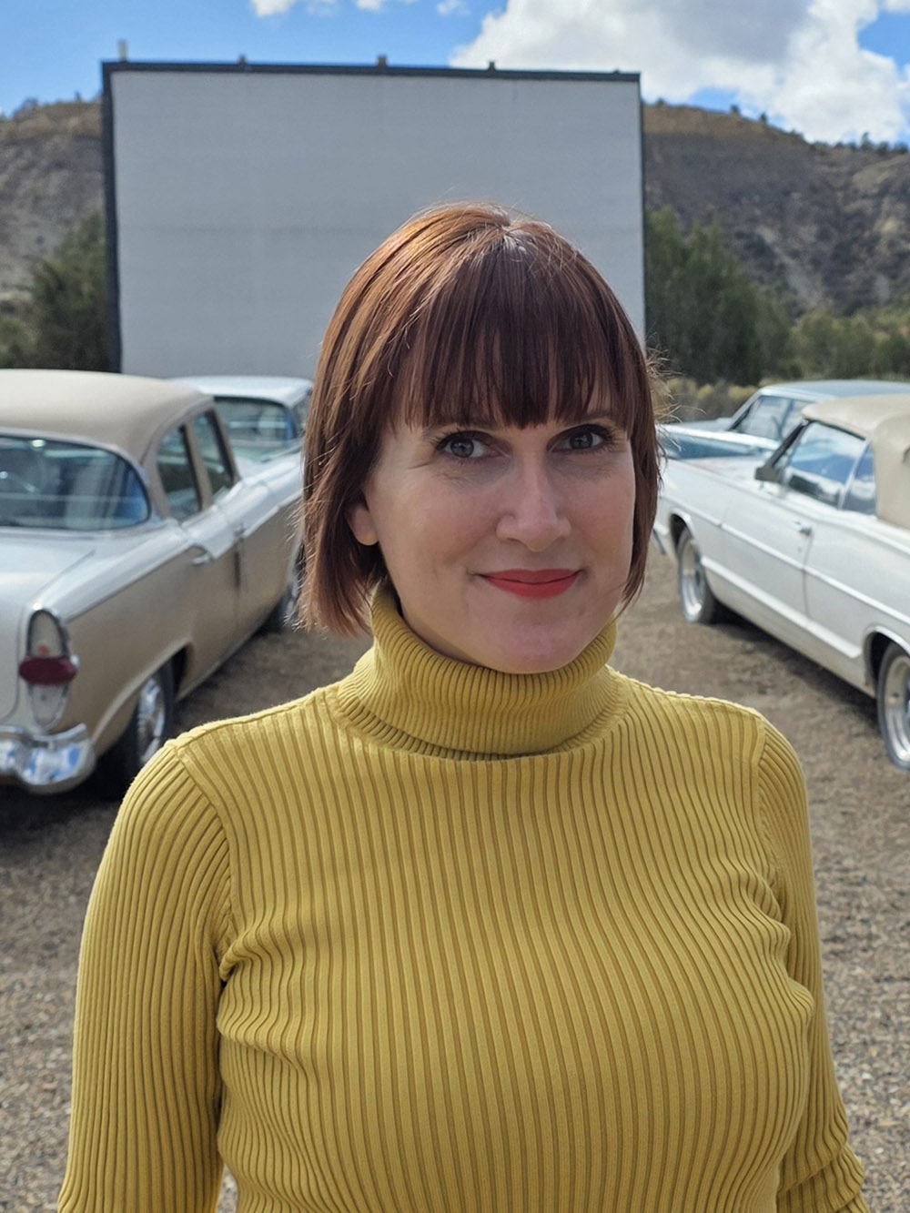 Lucy Jones, wearing a yellow ribbed turtleneck sweater, stands in the parking lot of a countryside drive-in movie theatre with retro and classic cars. Photograph by Mark Murray.