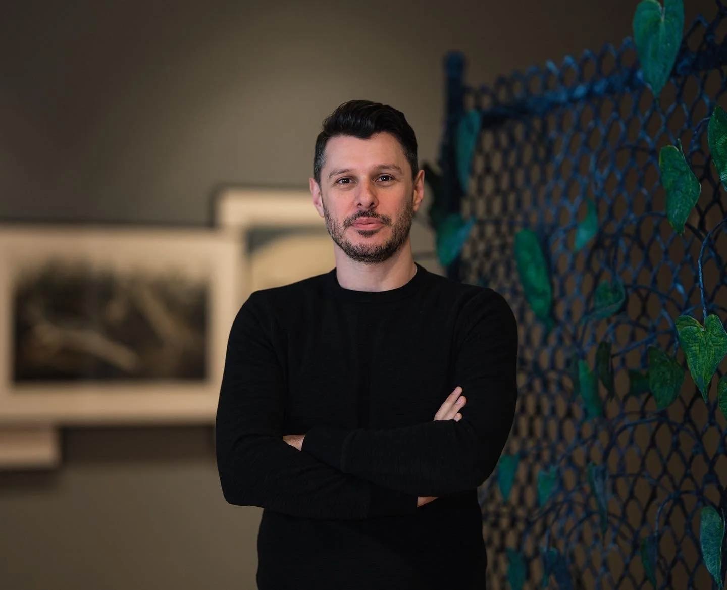 Aaron Garvey in black shirt with arms crossed standing in a gallery with artwork behind