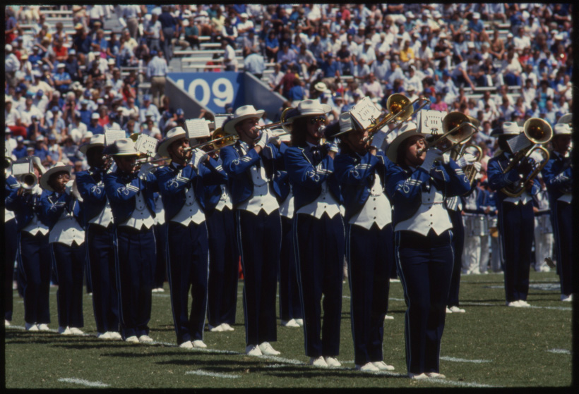 Color photo of the marching band while they perform in Commonwealth Stadium (now Kroger Field). A perfectly lined up section of trombonists of different heights with a section of Euphoniums right ot the right behind. The 2003 band uniform is a white brimmed hat, black and blue tailcoat with silver braided trim, a white waistcoat, black pants with blue side stripes, and white shoes.