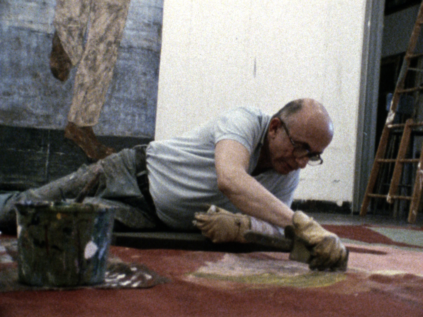 Still of Golub on the ground working on a piece of art wearing a grey tshirt and dark pants