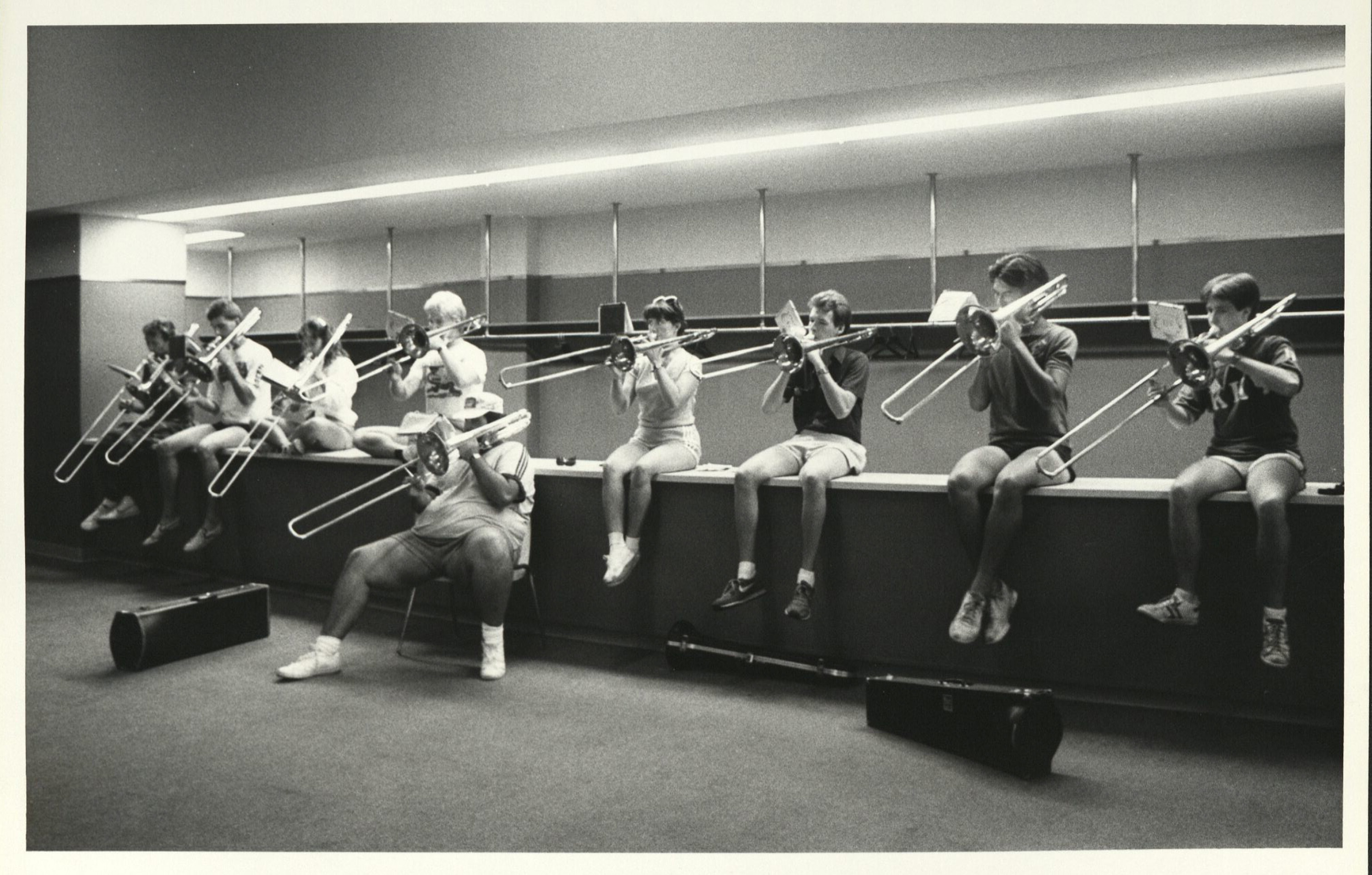 Black and white photo of 9 trombonists practicing together. Either of them site on top of a long counter across the back wall. A nineth, in the middle of your group, sits in a chair in front of them. A few empty trombone cases are scattered on the ground.