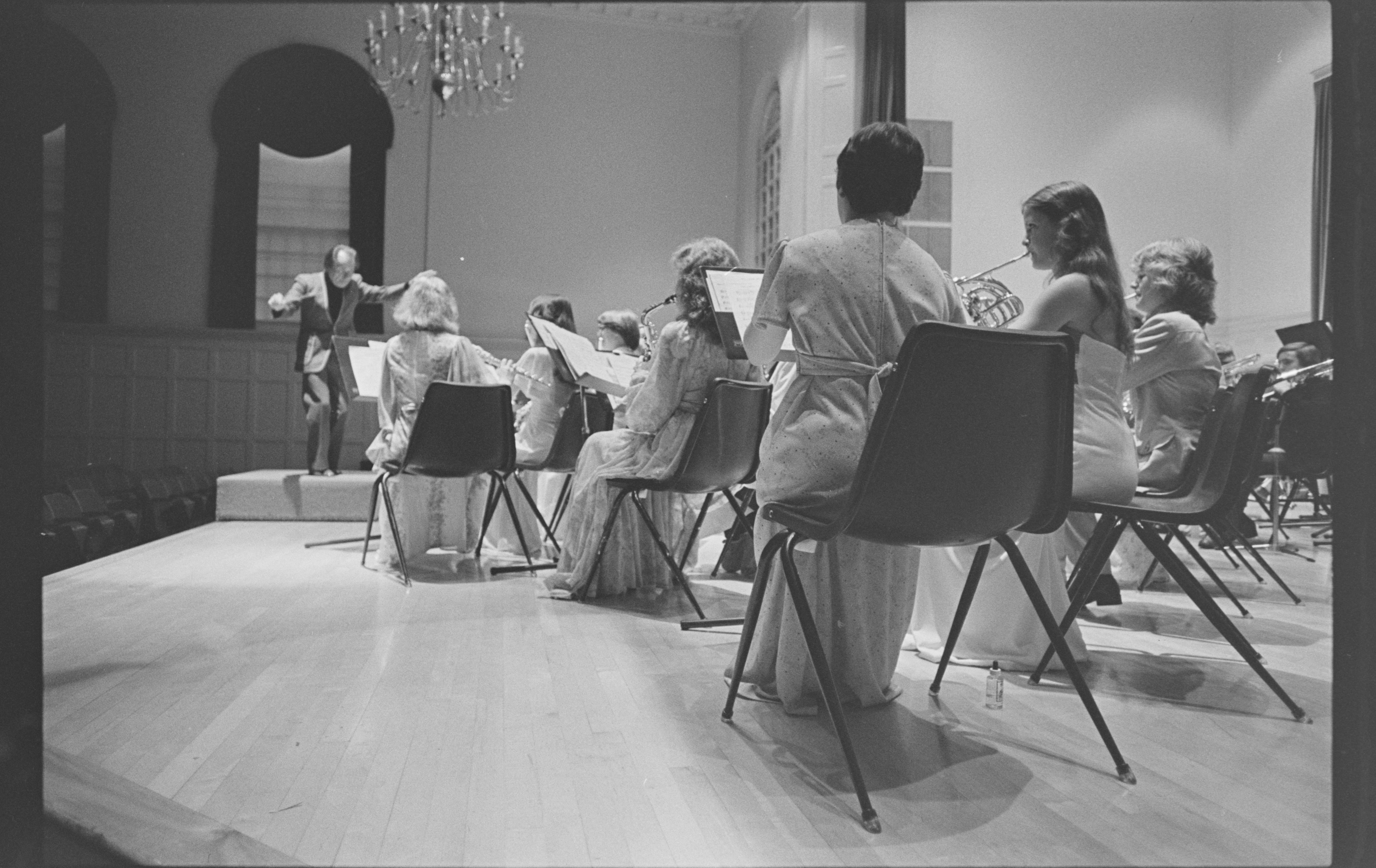 A black and white negative scan. The concert band comprised of brass and woodwind musicians plays in Memorial Hall. An energetic conductor can be seen in the distance. The women are in sparkly gowns and the men in suits.