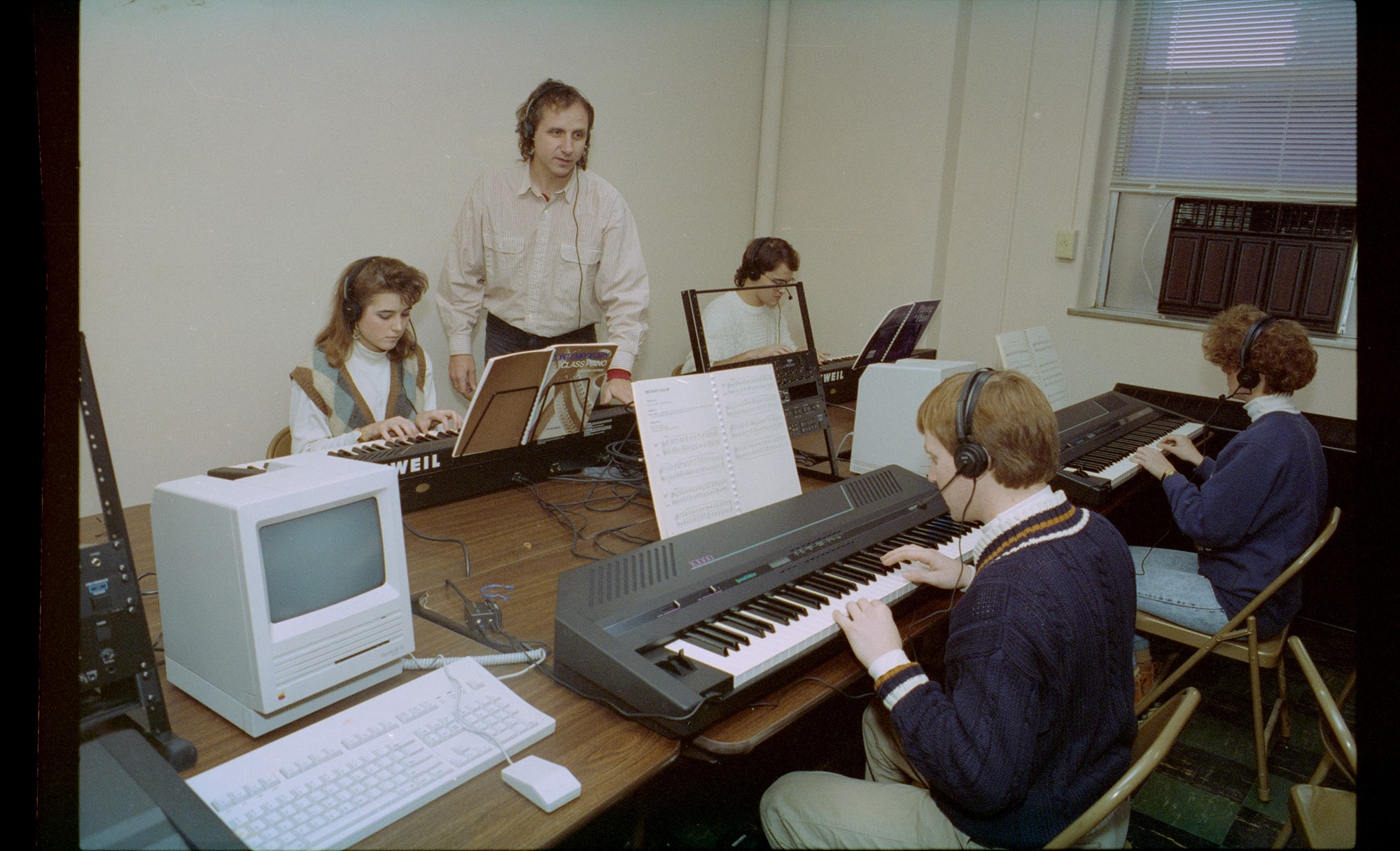 A color film scan. A chunky old computer with a tiny CRT screen sits on the near end of a long table. Four synthesizer keyboards are set up on the rest of the table, each with a student wearing wired headphones diligently practicing. A spaghetti of wires passes between them. An instructor stands in the background also wearing headphones and speaking to someone off camera.