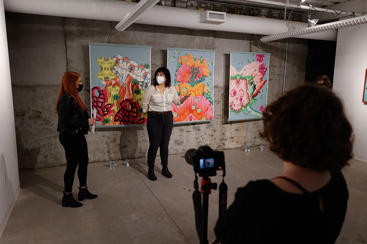 A masked graduate student in white button up shirt and black pants stands in front of very colorful painted wall scrolls with abstract floral designs hung from the ceiling in a stark industrial space. They gesture in explanation to an interviewer standing to the left. A person manning a camera can be seen to the right in the foreground.