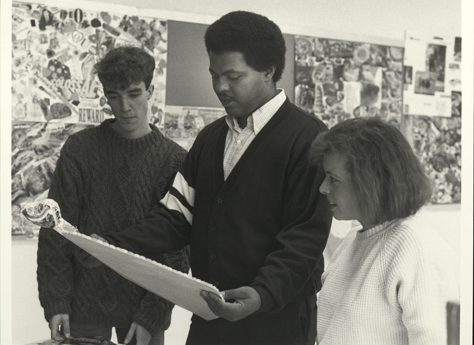 Black and white photo of three students standing together looking over an art piece held up by the man in the middle. The wall behind them is covered in sheets of sketches, mixed media word collages, photo prints, and more.