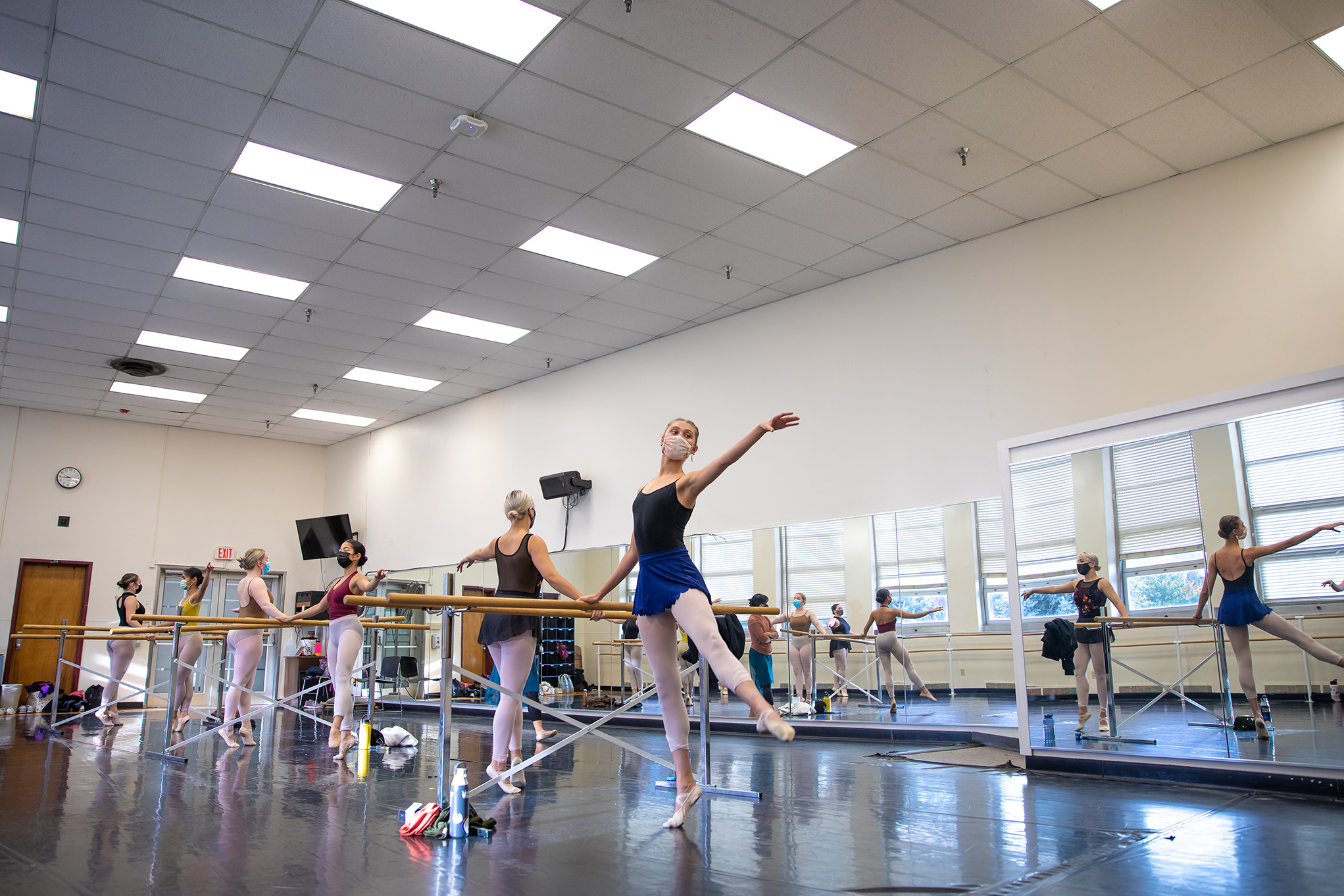 A spacious dance studio. Ballet bars are spaced extra wide in a single line through the space and dancers in masks practice their forms. A wall of the mirrors behind reflects them and the large windows of the opposite wall.