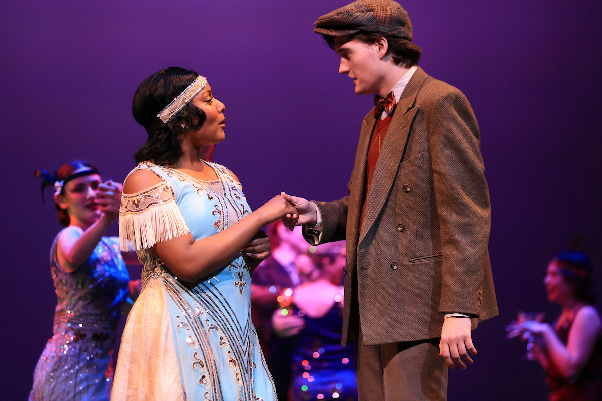 A woman in glamorously beaded 1920s dress converses with an enraptured man in a drab brown suit and cap. Other fancifully dressed ladies are partying in the background.