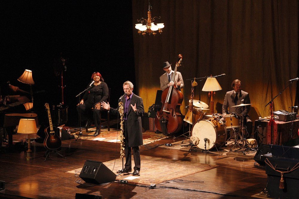 A softly lit stage with an ornate vintage light fixture hanging at the top of the frame. Hugh Laurie stands front and center by a decorated mic stand. Arranged behind him is a well dressed band made up of a backup singer, a bassist, and a drummer in view. Additional instruments and equipment to the right and left suggests additional musicians.