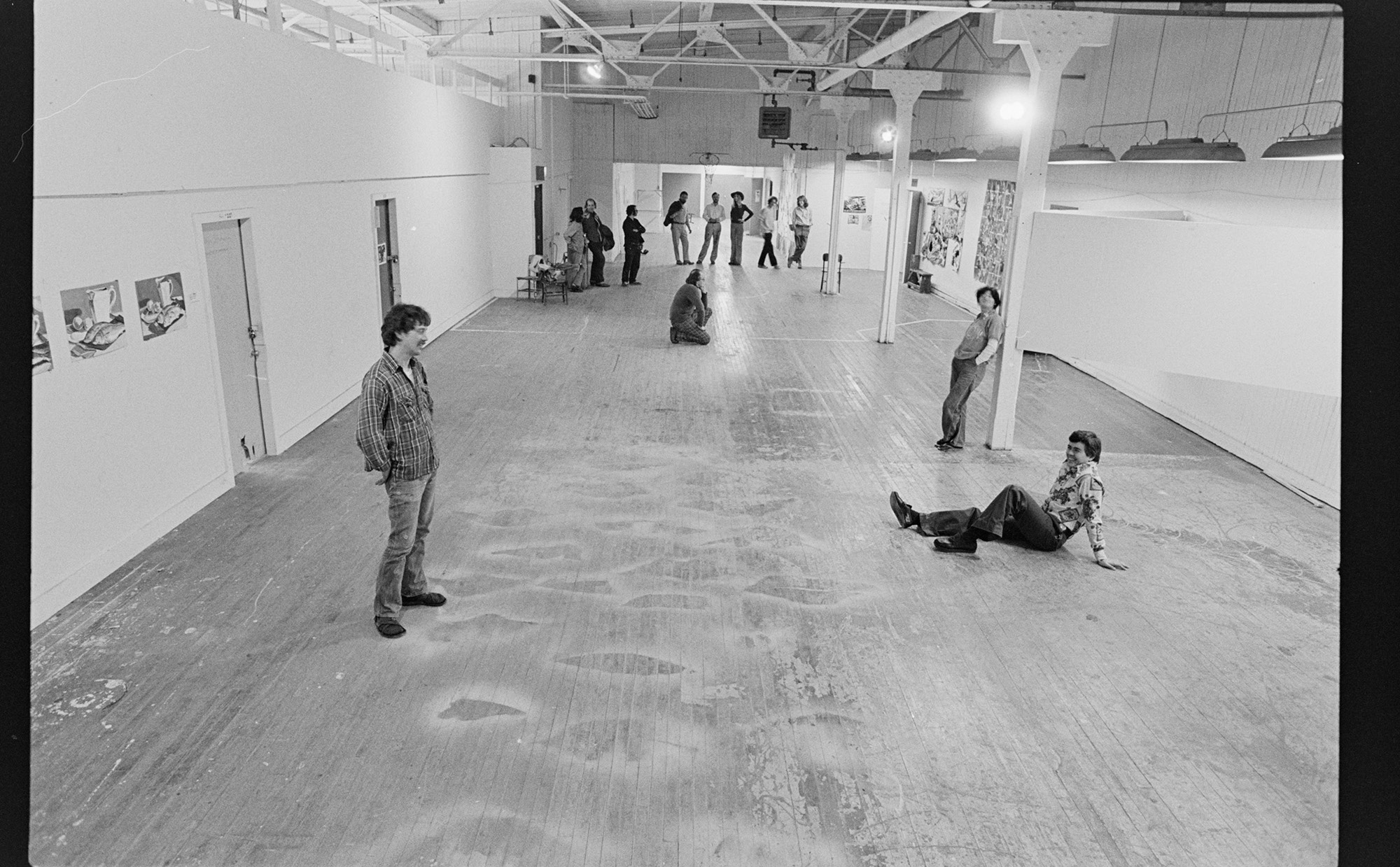 A black and white negative scan. A wide open white space with worn wooden floors has some art displayed in places on the walls and a few different gatherings of people. Some are students relaxing socially, others in deep contemplation of the art, and more others discussing future installations.