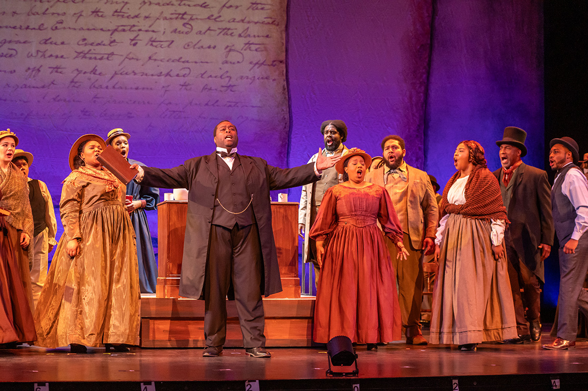 An ensemble of performers sing towards the sky, all dressed in historical costumes.