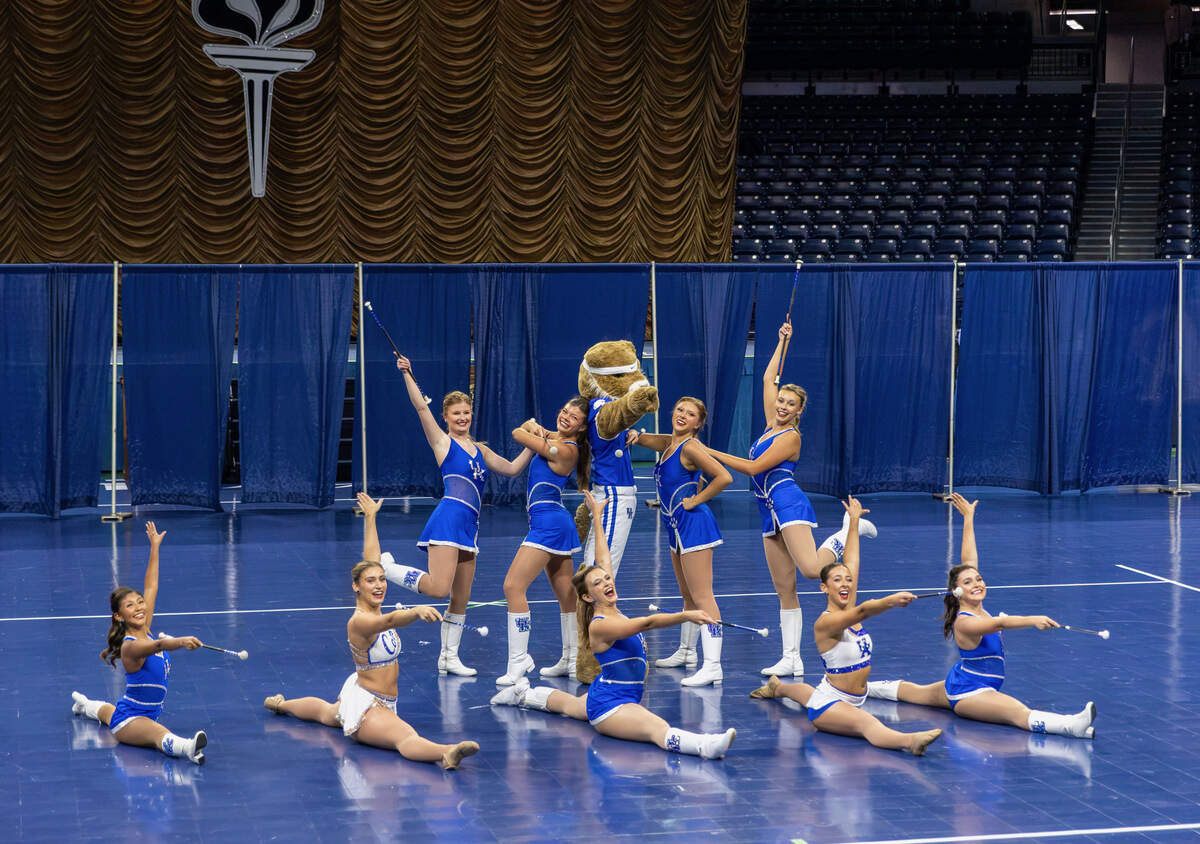 UK Feature Twirlers and Sweetheart Majorette Twirling line onstage wearing UK blue uniforms holding batons and posing for an end move with the UK Wildcat centerstage, against a blue backdrop of curtains.