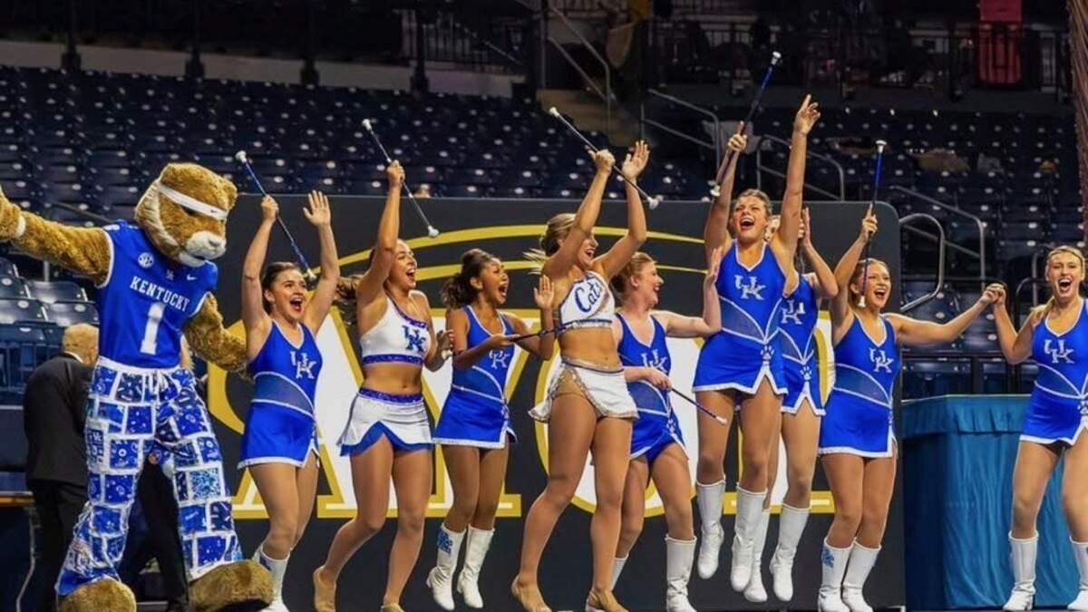 UK Feature Twirlers and Sweetheart Majorette Twirling line celebrates winning the national championship competition, wearing their UK blue uniforms jumping with arms raised next to the UK Wildcat in front of a crowd.