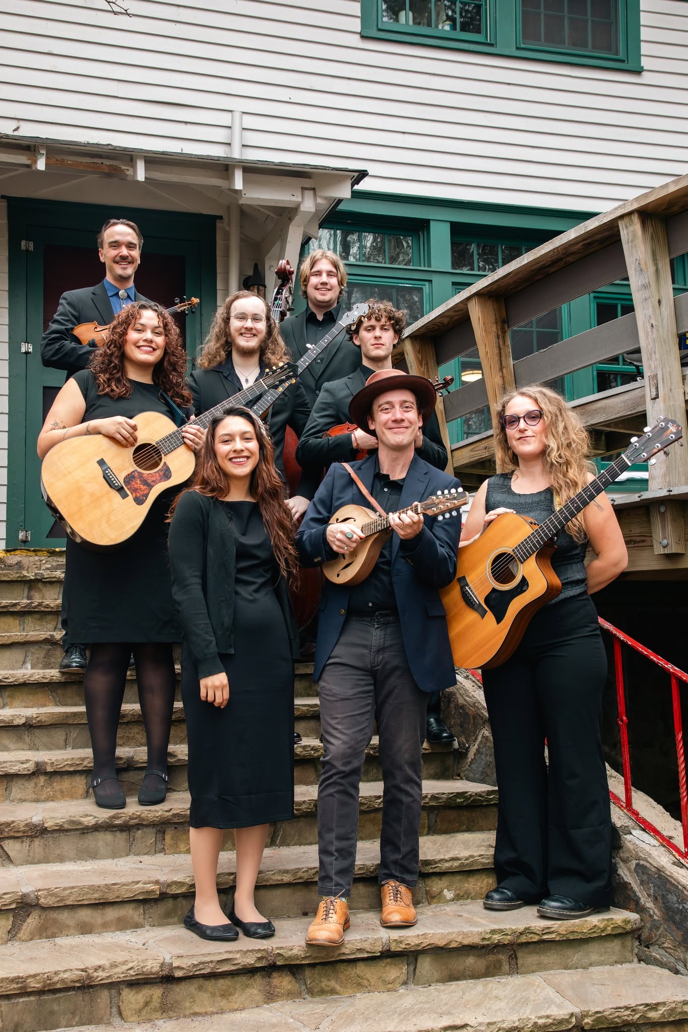 Eight members of the band stand together smiling with various instruments on rustic stone steps in front of a historic building.