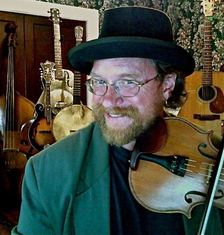 Craig Edwards in a green suit jacket and black hat greening while he holds a fiddle. A variety of common instruments in folk music are on display behind him.