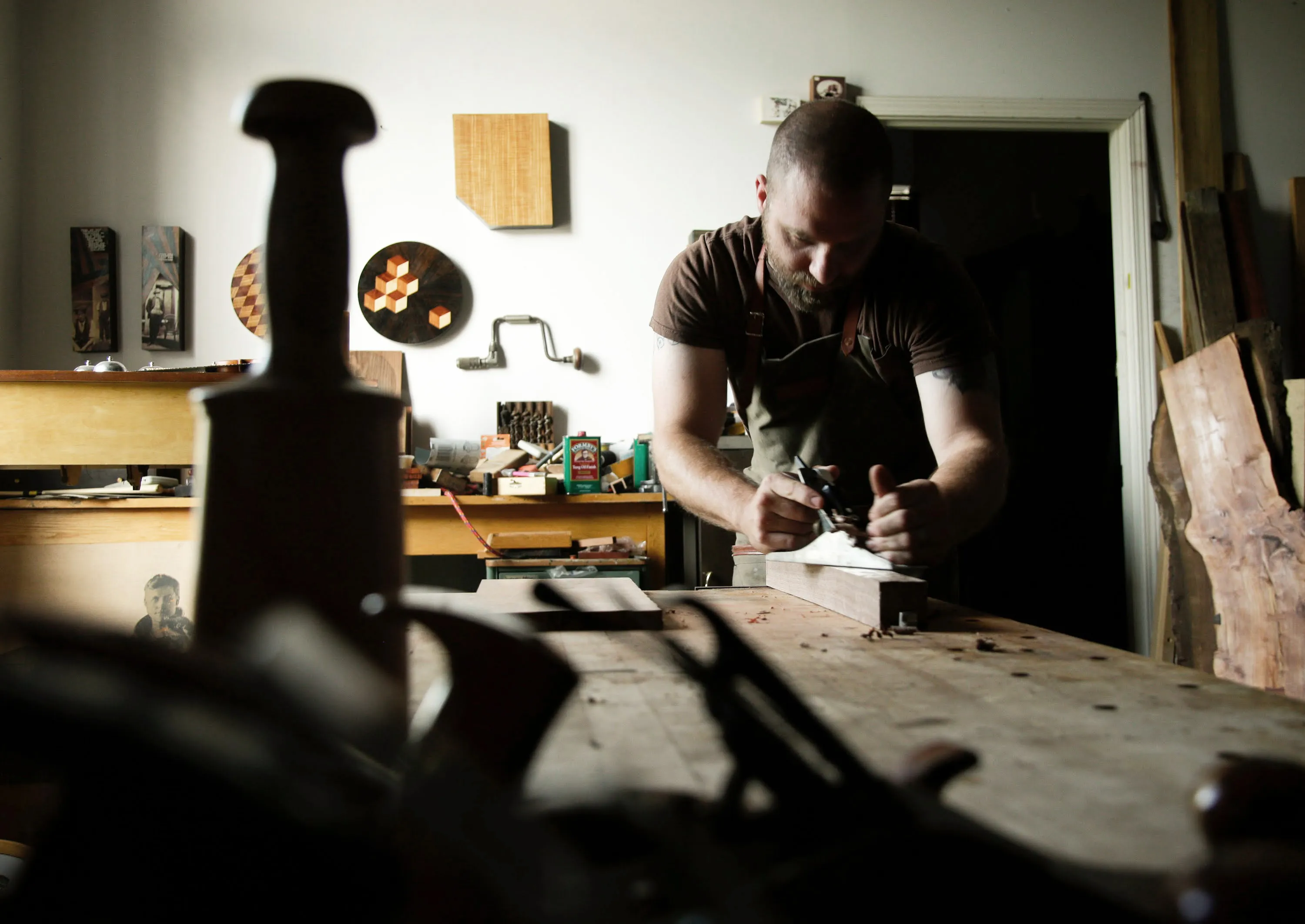 Daniel Graham in his workshop, hand planing a piece of wood. An assortment of tools, pieces in progress, and finished works can be seen around the room.