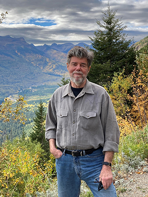 Jeff Warner stands on a mountain with a vibrant valley an other mountains spread out in the distance behind him.
