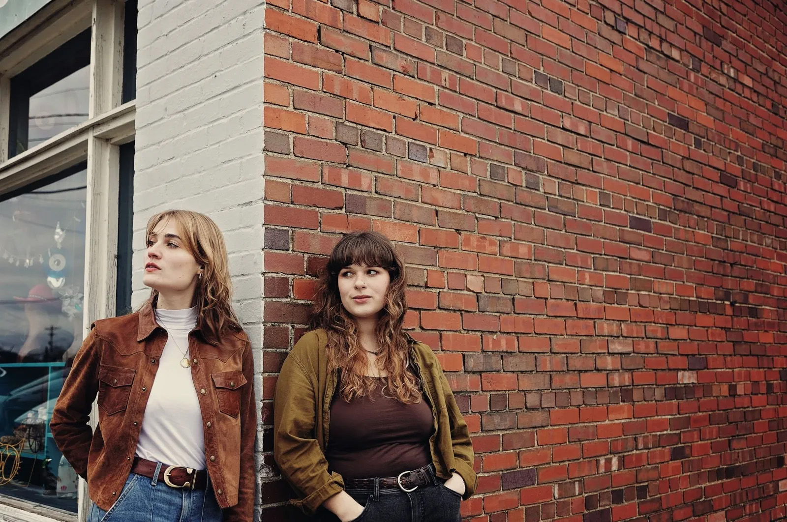 Two women look casually into the distance leaning against the corner of a small town brick andf glass storefront.