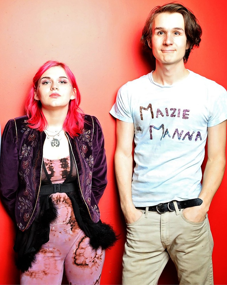 Two eclectic young starts stand in front of a vibrant grapefruit colored wall. Maizie Manna, with neon pink hair and dark jacket, stands on the left. A band mate with a shirt that reads "Maizie Manna" stands on the right.