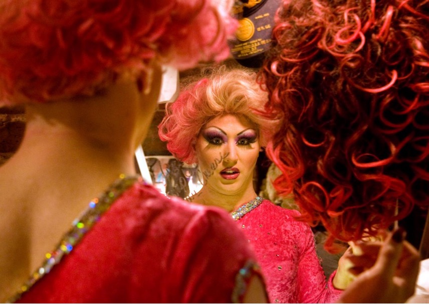 A performer in a bright pink costume and curly pink wig applies makeup while looking into a mirror backstage. The image is framed through the reflection, showing detailed stage makeup and costume embellishments. Another red-and-black curly wig appears in the foreground, partially surrounding the mirror. The warm lighting and dressing-room setting highlight preparations for a theatrical performance.