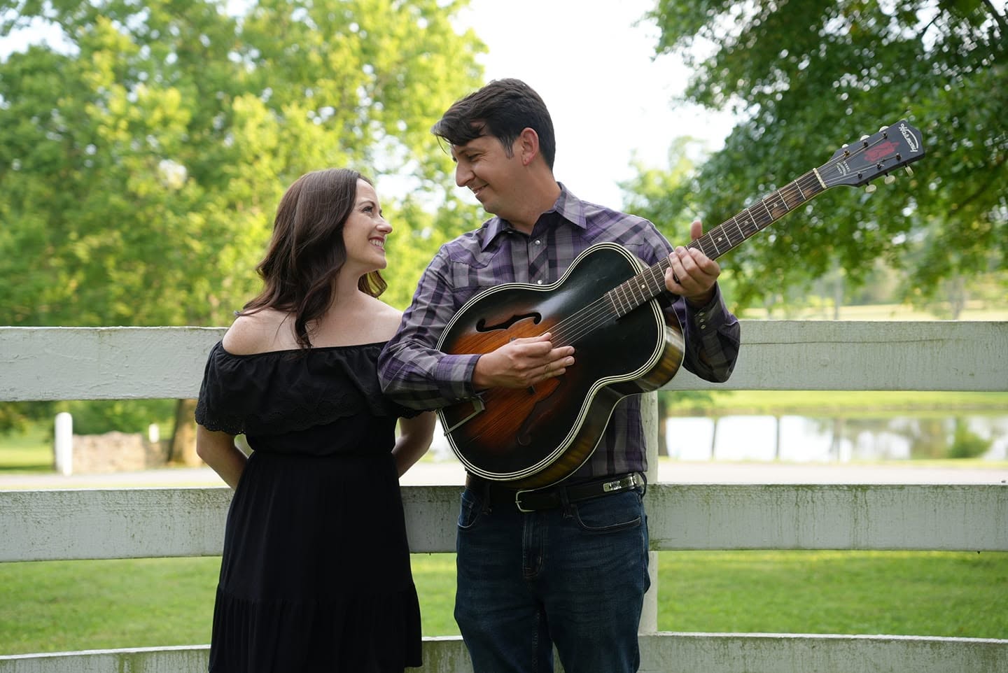 A woman and a man stand in front of a white farm fence with green grass and trees in the distance. The man has a guitar, and the two smile warmly at each other.