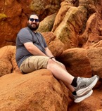 Joshua Cohen sitting on a large orange sandstone boulder in the wilderness.