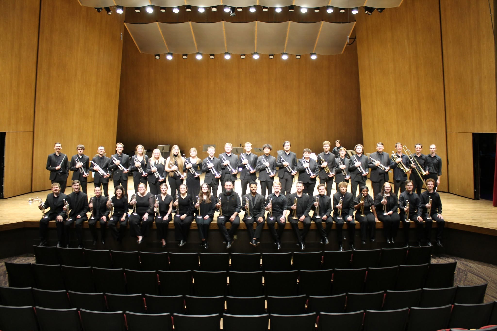 39 students plus Professor Jason Dovel are in black formal wear on the softly lit Singletary Recital Hall stage. Half of them are standing in a row behind the other half who are sitting on the edge of the stage.