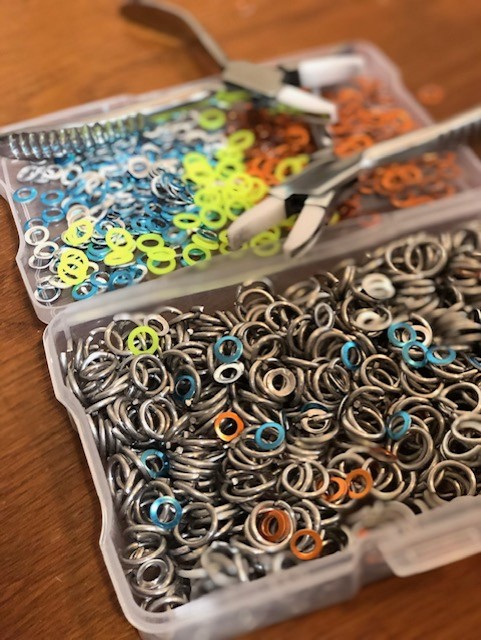 A an open clear plastic storage box sitting on a wooden table holds multicolor metal rings. A couple sets of jeweler's pliers sit on top.