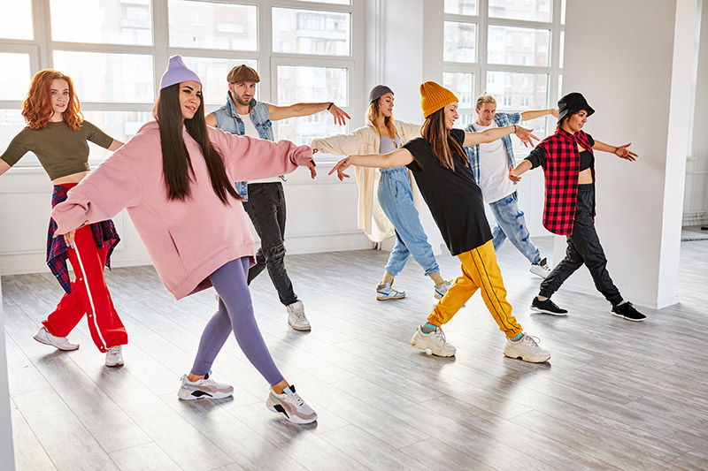 A group of dancers in colorful street-like fashions strike a pose in a bright dance studio.