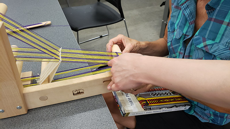 A student sits with a freshly warped inkle loom and a pattern booklet in their lap.