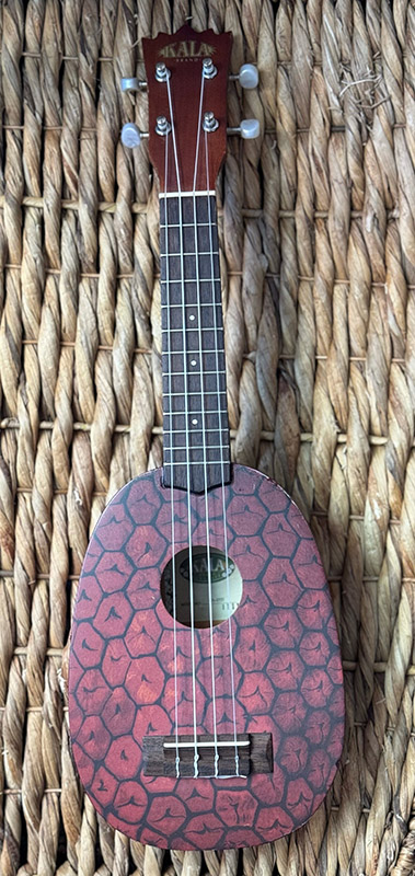 A ukulele with a pineapple skin print on the body of instrument, laying on a woven grass mat.