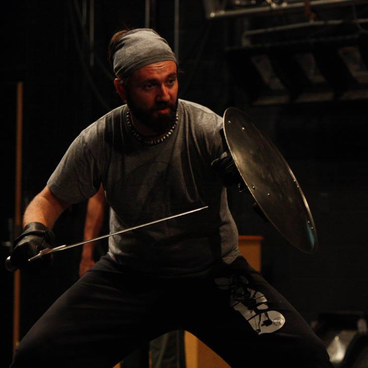 Instructor Andrew Ray takes a stance with a sword and shield on a dark stage.