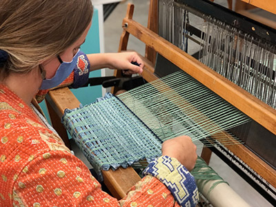 A student using an assortment of yarn and fabric pieces to weave on a floor loom.