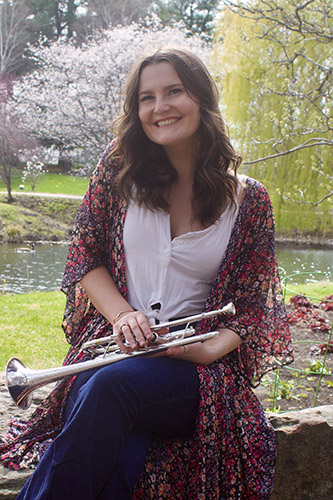 Julia Stowell sits on a low stone fence in a blooming botanical garden with a pond behind her. In her lap is a silver trumpet.