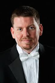 Headshot of Jim Daughters, wearing a tuxedo with a whie bowtie in front of a black background.