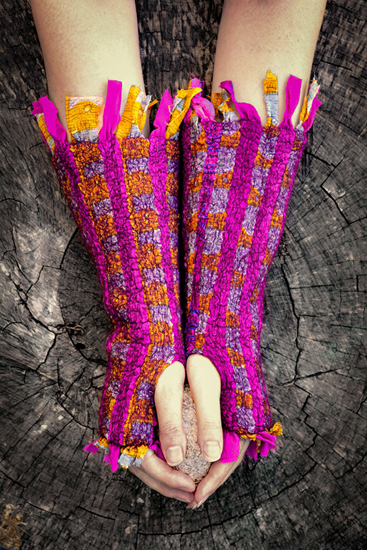 Extremely colorful magenta, purple, and orange chequered arm warmers adorn a person's hands and forearms while leaned against a weathered tree stump and holding a rock between both hands.