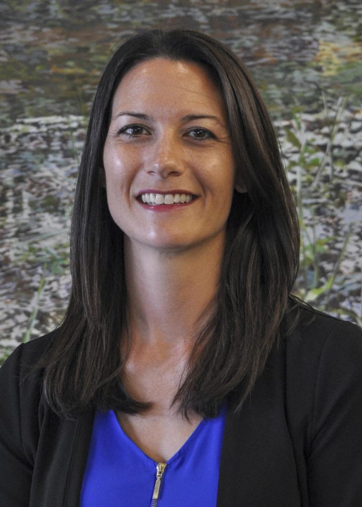 Headshot of Erin Brodnar, smiling and wearing a blue shirt with a black cardigan, standing in front of an abstract painted background.