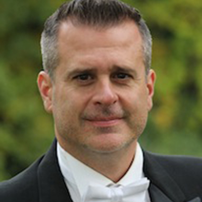 Headshot of Mike Fansler, wearing a tuxedo with a white bowtie outdoors in front of blurry distant greenery.